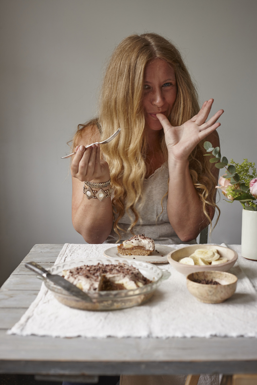 Joanna Henderson Food photography. Hanna eating banoffee pie seated at a wooden table with bananas and cocoa and fresh flowers